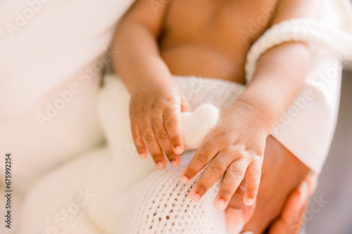 Newborn baby is held in his parents' arms as he holds a heart with his hands. Close-up of his fingers 