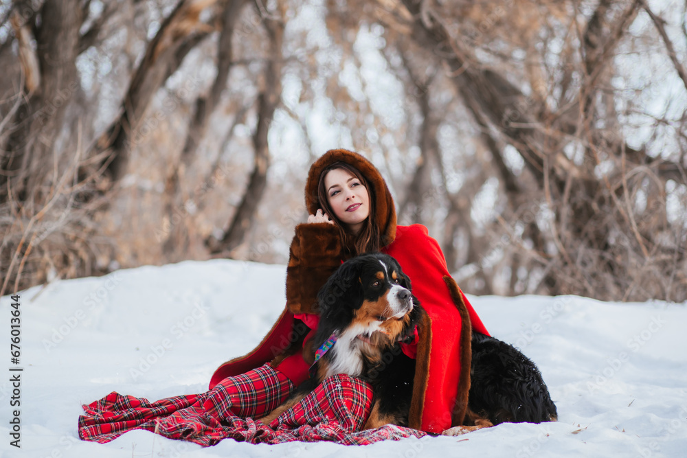 woman in winter in an ancient red coat and plaid skirt in winter in the snow with a dog of the Bernese Mountain Dog breed. Fairytale image of Little Red Riding Hood with a big dog.