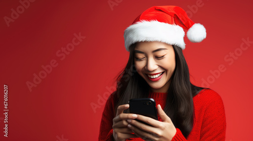 A young woman in a sweater and Santa hat is happily looking at her smartphone against a red background.