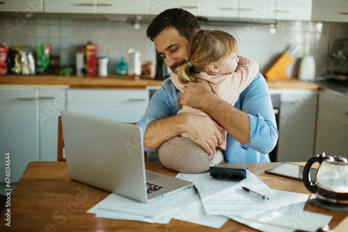 Little young daughter hugging father in kitchen with documents on desk photo