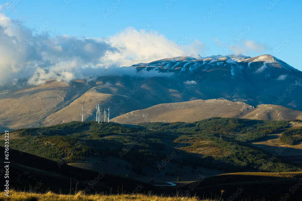 Naklejka premium Wind farm with turbines on the mountains. Eco-sustainable industry with nature-friendly technologies for clean energy in a better environment. Conceptual mountainous landscape