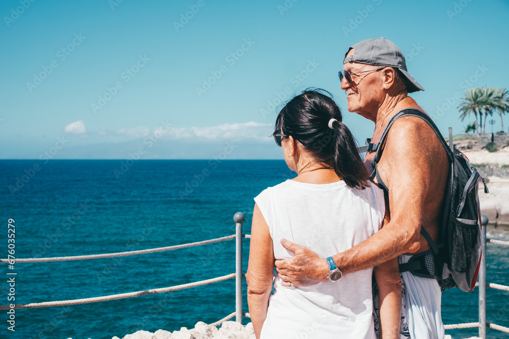 Back view of caucasian senior couple hugging outdoors in sea vacation looking at horizon over water. Elderly people enjoying healthy lifestyle  retirement and travel
