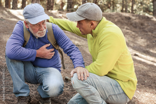Old senior man hiking in the mountains with his young nephew pauses to catch his breath, touching his heart. Elderly grandfather feeling unwell and his grandson assists him