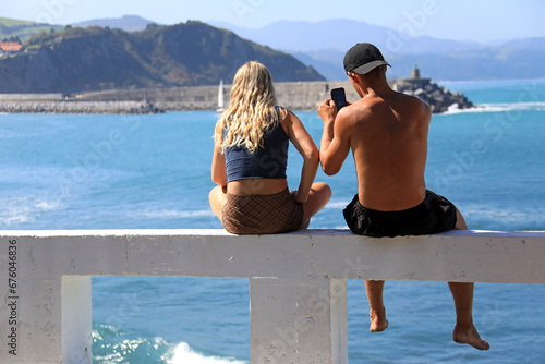 jovenes pareja sentados mirando al mar con móvil desde un pretil zumaia verano getaria país vasco 4M0A2550-as23 photo