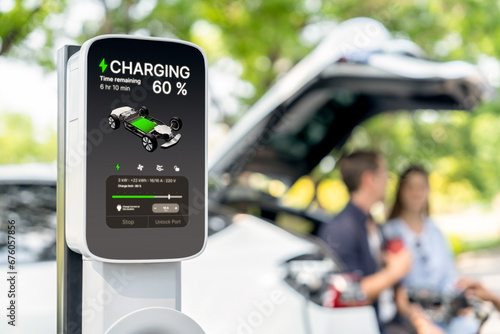Focused EV charging station monitor display battery status for electric car on blurred background of couple sitting on car's trunk while recharging battery during natural travel and holiday. Exalt
