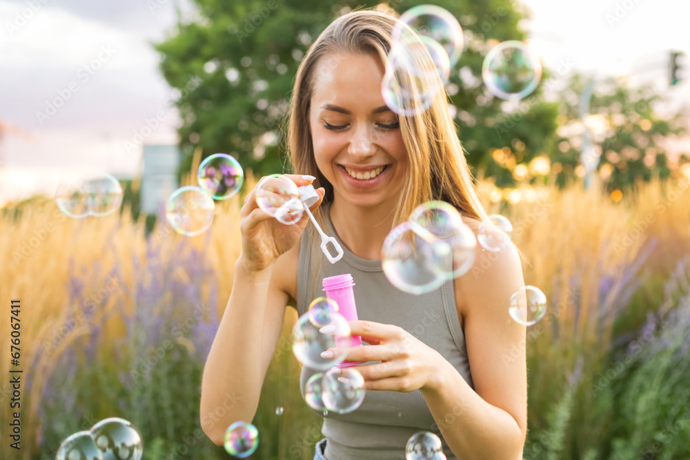 Happy young woman with long loose hair blows soap bubbles laughing in city garden. Smiling lady blows bubbles having fun in park in evening