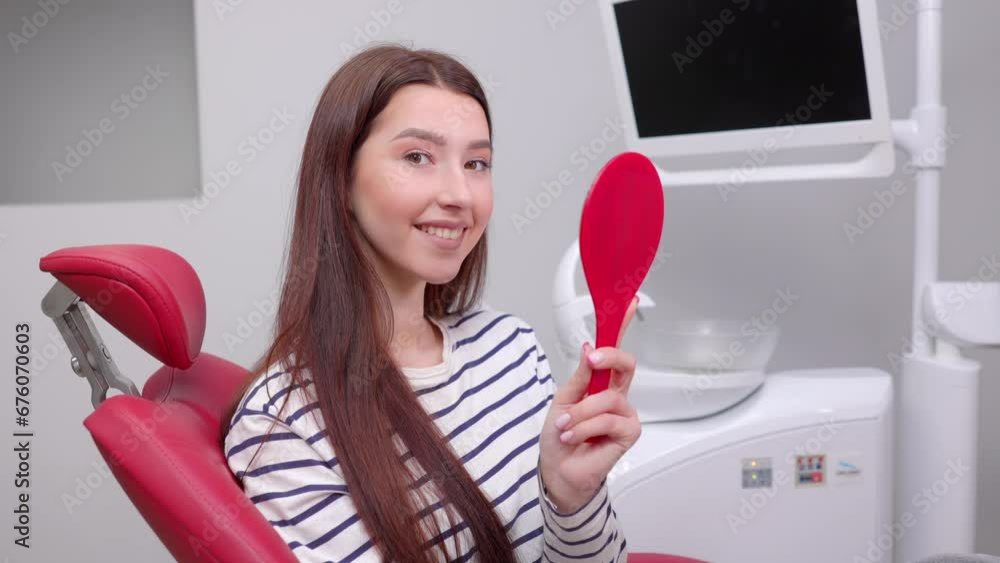 Happy caucasian female patient looking at mirror after dental treatment in clinic. Cheerful woman sitting in chair in dental cabinet and enjoying her beautiful smile Happy caucasian female patient looking at mirror after dental treatment in clinic. Cheerful woman sitting in chair in dental cabinet and enjoying her beautiful smile