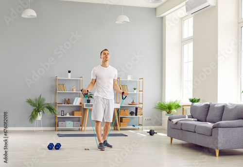 Young sporty man in homewear doing stretching exercises with rubber band in the living room at home. Athletic guy enjoy exercising. Fitness, workout sport and home training concept. photo