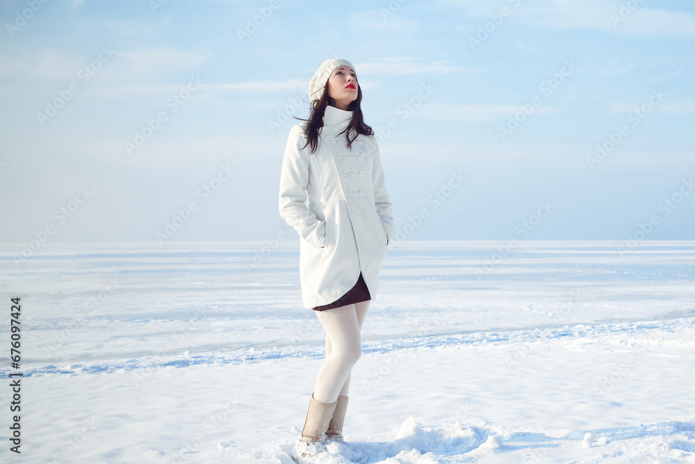 Winter fashion concept. Full length portrait of fashionable model in white coat and beret standing at the snow seaside. Sunny weather. French style. Text space. Outdoor shot. Winter fashion concept. Full length portrait of fashionable model in white coat and beret standing at the snow seaside. Sunny weather. French style. Text space. Outdoor shot.