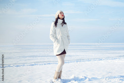 Winter fashion concept. Full length portrait of fashionable model in white coat and beret standing at the snow seaside. Sunny weather. French style. Text space. Outdoor shot.