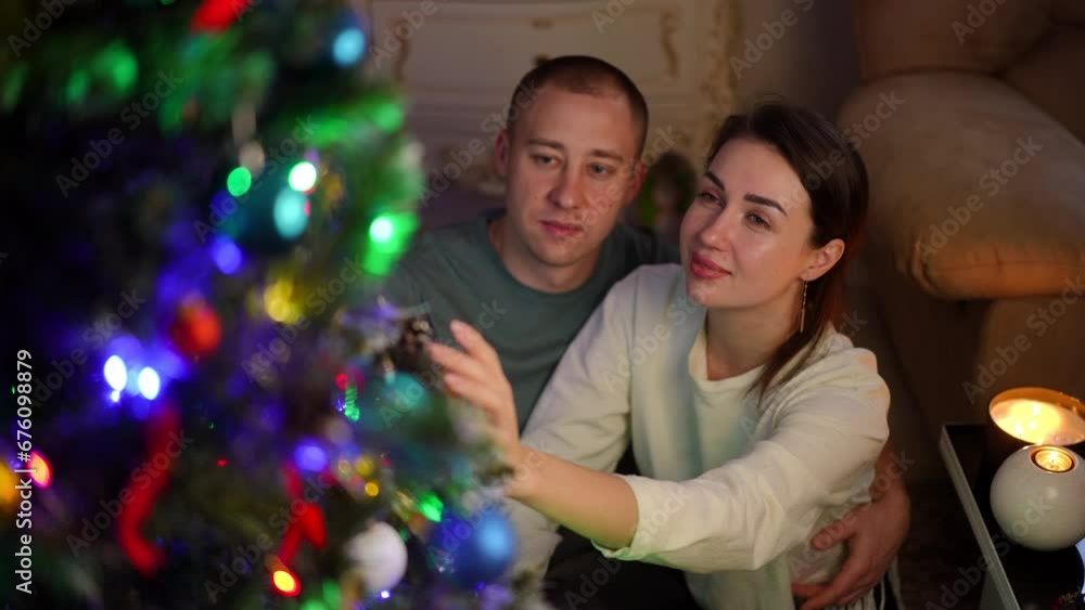 Slow motion. A couple in love enjoys the view of the Christmas tree. The woman shows the man a beautiful Christmas tree decoration on the Christmas tree, gently touching it Slow motion. A couple in love enjoys the view of the Christmas tree. The woman shows the man a beautiful Christmas tree decoration on the Christmas tree, gently touching it