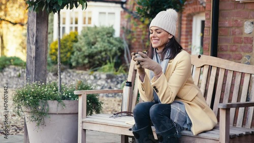 Woman dressed for autumn or winter walk outside house sitting on bench with flask of hot drink - shot in slow motion photo
