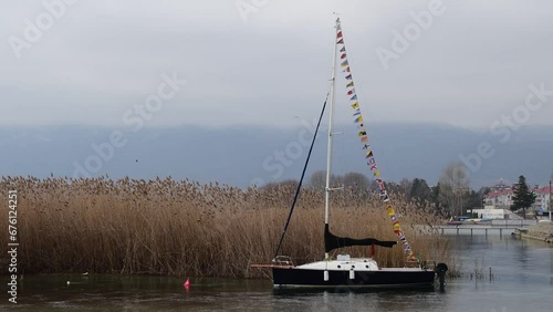 Boats sway in the waters of Lake Ohrid, tied to thick reeds. Flags are flying on the mast.
