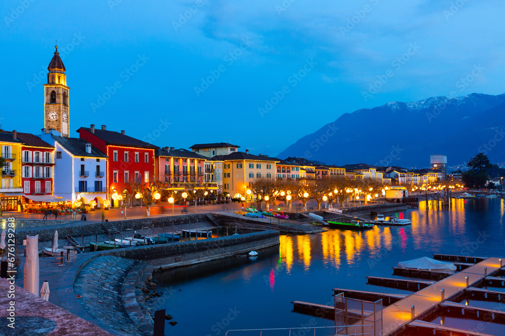 View of embankment in Ascona, canton of Ticino, Switzerland. Houses along waterfront, turned on city lights in evening.