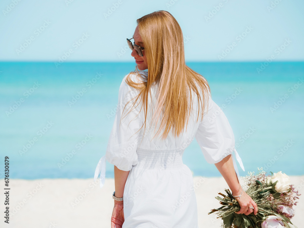 Joyful Young Woman Holding a Bouquet of Flowers on a Beach with Blue Sky Background