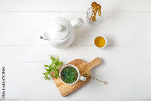 Teapot with cup of fresh mint tea, honey and spoons on white wooden background