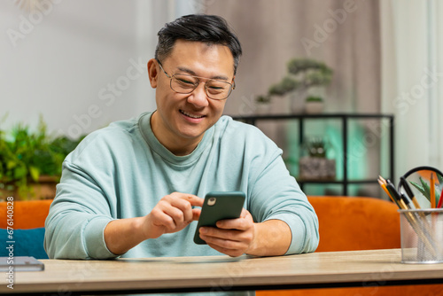 Asian man sits on couch uses mobile phone smiles at home room apartment. Middle-aged Chinese guy texting share messages content on smartphone social media applications online watching relax movie photo