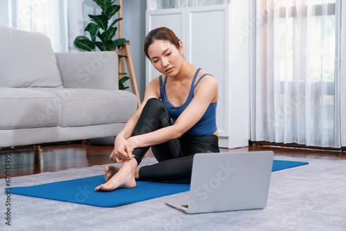 Asian woman in sportswear sitting on the house floor while watching online exercise training video on laptop. Attractive girl engage in her pursuit of healthy lifestyle. Vigorous photo