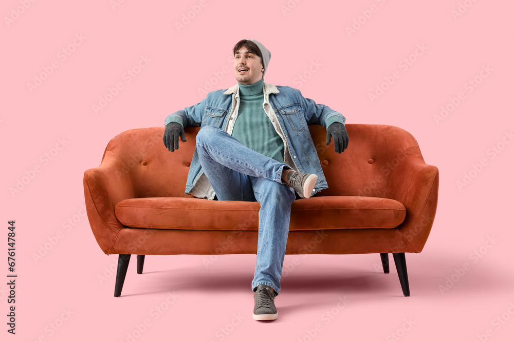Young man in winter clothes sitting on red sofa against pink background Young man in winter clothes sitting on red sofa against pink background