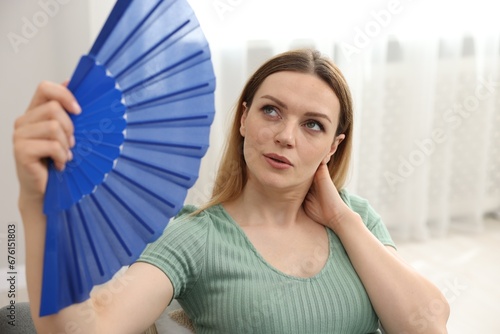 Woman waving blue hand fan to cool herself on sofa at home