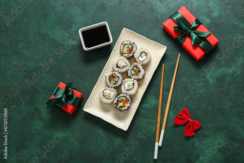 Plate with tasty sushi rolls, soy sauce, gift boxes and bow for Christmas party on green background