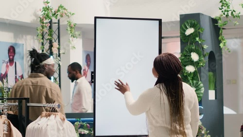 African american woman in fashion boutique using mockup augmented reality screen to look at clothes options to try on. Customer using led kiosk to visualize outfit combinations in store before buying photo