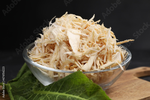Grated horseradish in bowl, leaf and board on black table, closeup