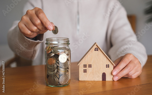 Closeup image of a woman putting coins into a glass jar while holding a wooden house model for saving money concept