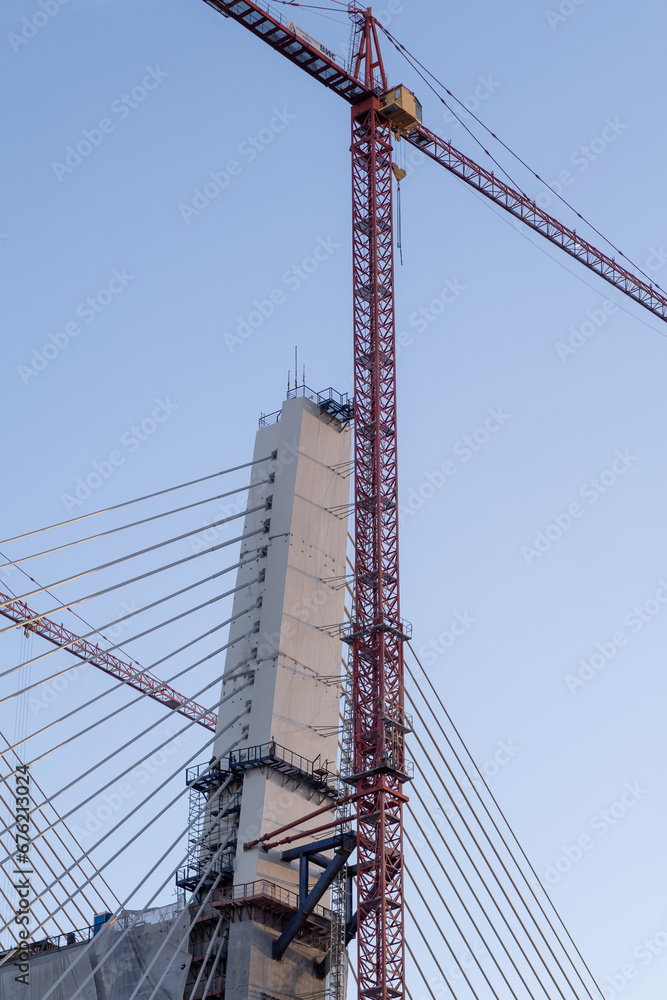 Crane and bridge construction against a blue sky background. Builders work on large construction sites, and there are many cranes working. There are a lot of cables on the bridge Crane and bridge construction against a blue sky background. Builders work on large construction sites, and there are many cranes working. There are a lot of cables on the bridge