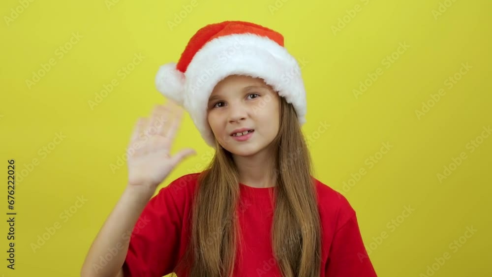 Cute smiling little girl in Santa hat waving hand, posing isolated over yellow color background wall in studio. Cute smiling little girl in Santa hat waving hand, posing isolated over yellow color background wall in studio.