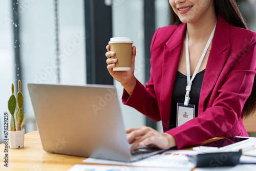 Confident Asian businesswoman holding a paper coffee cup drinking coffee while taking a break at her desk working on corporate financial business documents on a laptop computer.