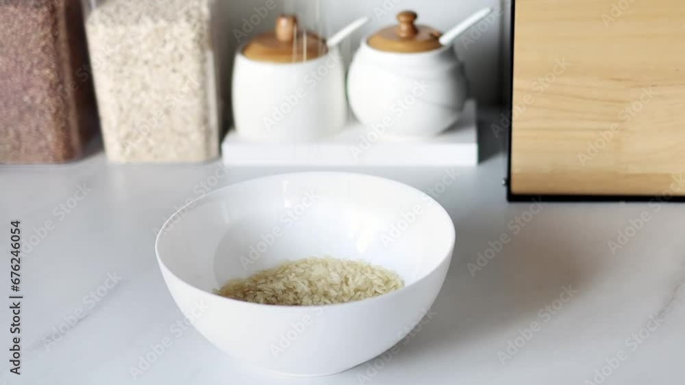 White raw rice is poured into a white plate against the background of a modern kitchen in slow motion. Healthy food and nutrition concept. Safe, ecological, and healthy products. White raw rice is poured into a white plate against the background of a modern kitchen in slow motion. Healthy food and nutrition concept. Safe, ecological, and healthy products.
