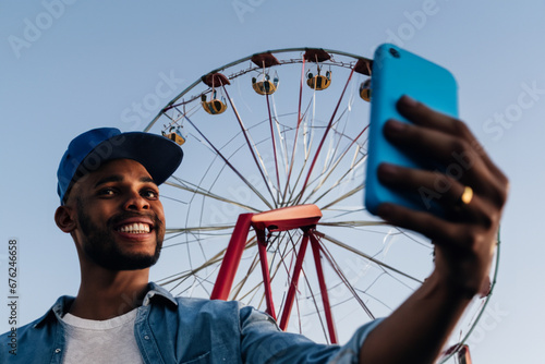 guy with a Ferris wheel. a young guy in a cap and blue shirt takes a selfie on his phone against the backdrop of the Ferris wheel attraction