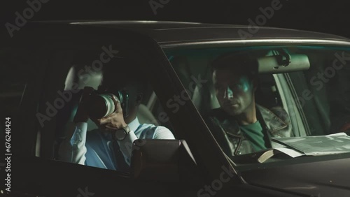 Waist up of two multiethnic male cops stalking and tracing down criminal in car at night. Black male officer taking photos on professional camera and showing them to colleague at driver seat photo