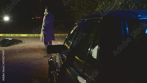 Back view full shot of African American male detective in long camel trench coat crossing yellow barrier tape and examining murder scene at night. Corpse in black plastic bag lying on ground photo