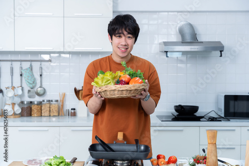 Happy portrait of asian young man holding a basket of vegetables of standing a cheerful preparing food and enjoy cook cooking with vegetables, while standing on a kitchen Condo life or home