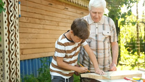 An elderly gray-haired man teaches boys carpentry outdoors. Grandfather teaches his grandchildren to carpenter. A boy draws a pencil line on a wooden board.