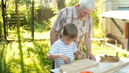 An elderly gray-haired man teaches boys carpentry outdoors. Grandfather teaches his grandchildren to carpenter. A boy measures a wooden board with a tape measure.