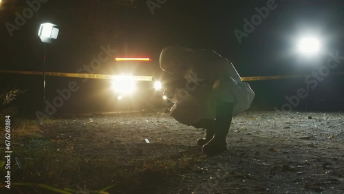 Anonymous forensic expert wearing protective coveralls, respirator mask and gloves taking photographs of crime scene on camera while working at night photo