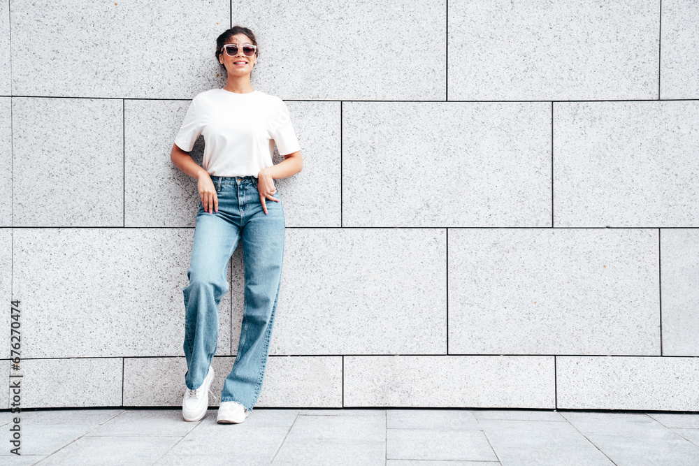 Young beautiful smiling hipster woman in trendy summer white t-shirt and jeans clothes. Carefree woman, posing in the street at sunny day. Positive model outdoors near wall. In sunglasses Young beautiful smiling hipster woman in trendy summer white t-shirt and jeans clothes. Carefree woman, posing in the street at sunny day. Positive model outdoors near wall. In sunglasses
