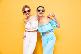 Two young beautiful smiling brunette hipster female in trendy summer clothes. Sexy carefree women posing near yellow wall in studio. Positive models having fun. Cheerful and happy. In sunglasses