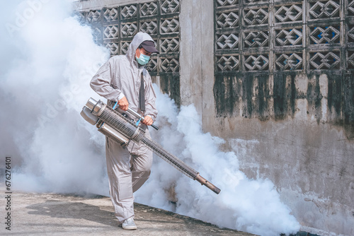 Healthcare worker using Fogging Machine Spraying chemical to eliminate Mosquitoes and prevent Dengue fever in drainage ditch near the old fence wall, full length view photo