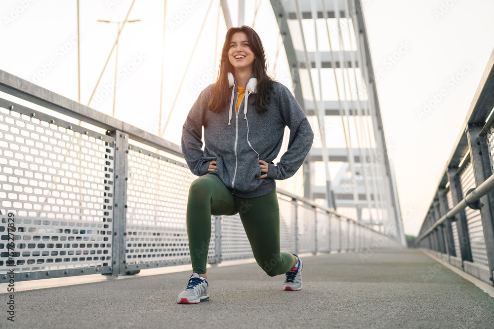 Fototapeta premium Young sportswoman warming up on a bridge while smiling in the morning. Smiling fitness girl doing lunge exercise while holding hands on her waist.