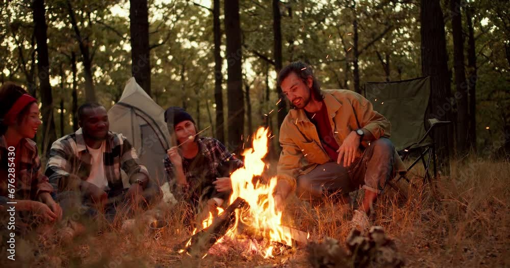 A brunette man in a light green jacket adjusts firewood in a fire while other participants in a group hike sit and enjoy the views of bright flames, fry marshmallows and have fun in the forest A brunette man in a light green jacket adjusts firewood in a fire while other participants in a group hike sit and enjoy the views of bright flames, fry marshmallows and have fun in the forest