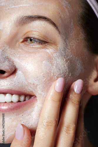 Dermatology. Exfoliating. Skin care facial scrub. Beauty portrait of young happy woman is applying white peeling cosmetic product to whole her face. Daily skincare routine. Dermatology. Exfoliating photo