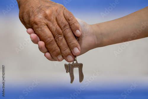 Am Israel Chai. Hands of an elderly man holding the hand of a child close up. they holding a silver Chai key chain, which spell "life" in hebrew. Blue and White background, Israel flag