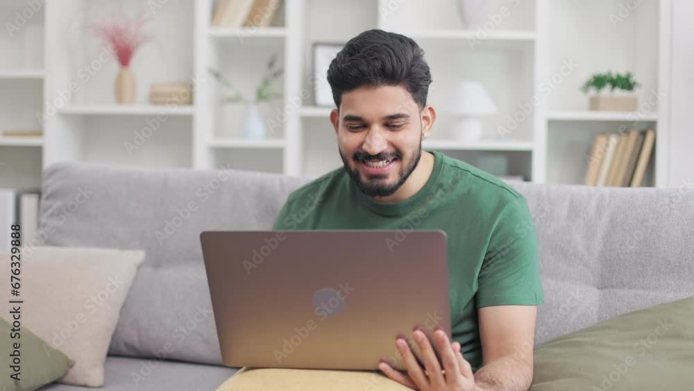 Cheerful indian guy dressed in casual clothes using wireless laptop for pleasant video conversation at home. Handsome bearded man sitting on couch, waving with hand and sincerely smiling. Cheerful indian guy dressed in casual clothes using wireless laptop for pleasant video conversation at home. Handsome bearded man sitting on couch, waving with hand and sincerely smiling.