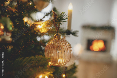 Merry Christmas! Stylish christmas gold bauble on tree close up against burning fireplace. Beautiful decorated christmas tree with vintage ornaments, ribbons and lights. Christmas background. photo