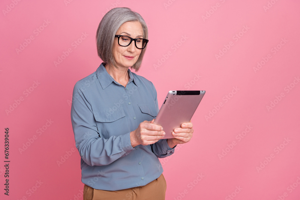 Photo of focused smart woman with gray hairstyle dressed blue stylish shirt in eyewear read news on tablet isolated on pink background Photo of focused smart woman with gray hairstyle dressed blue stylish shirt in eyewear read news on tablet isolated on pink background