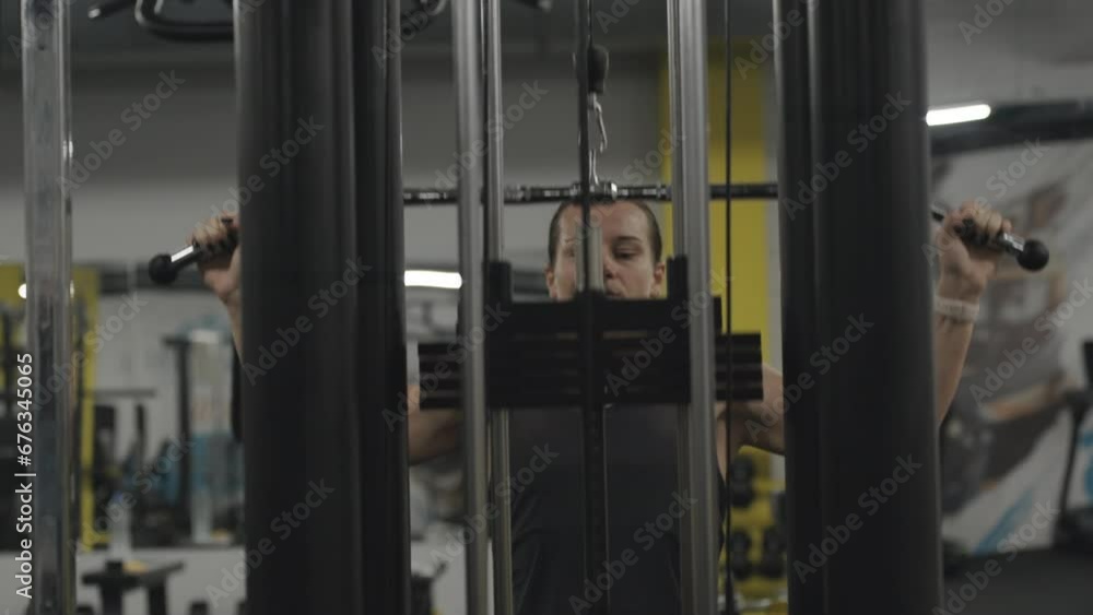A woman in a cable machine performs seated lat pulldowns, developing her back muscles. Viewed from the front through the machine. A woman in a cable machine performs seated lat pulldowns, developing her back muscles. Viewed from the front through the machine.